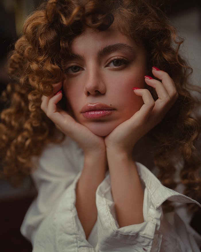 Close-up portrait of a woman with curly hair and white shirt, resting her chin on hands.