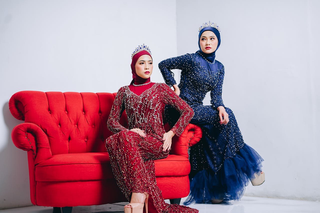 Two women in elegant hijabs and gowns with crowns sitting on a red sofa in a studio.
